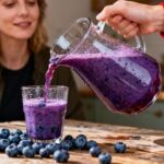 Jus de bleuet vibrant versé dans un verre par une main féminine, avec des bleuets frais sur une table en bois